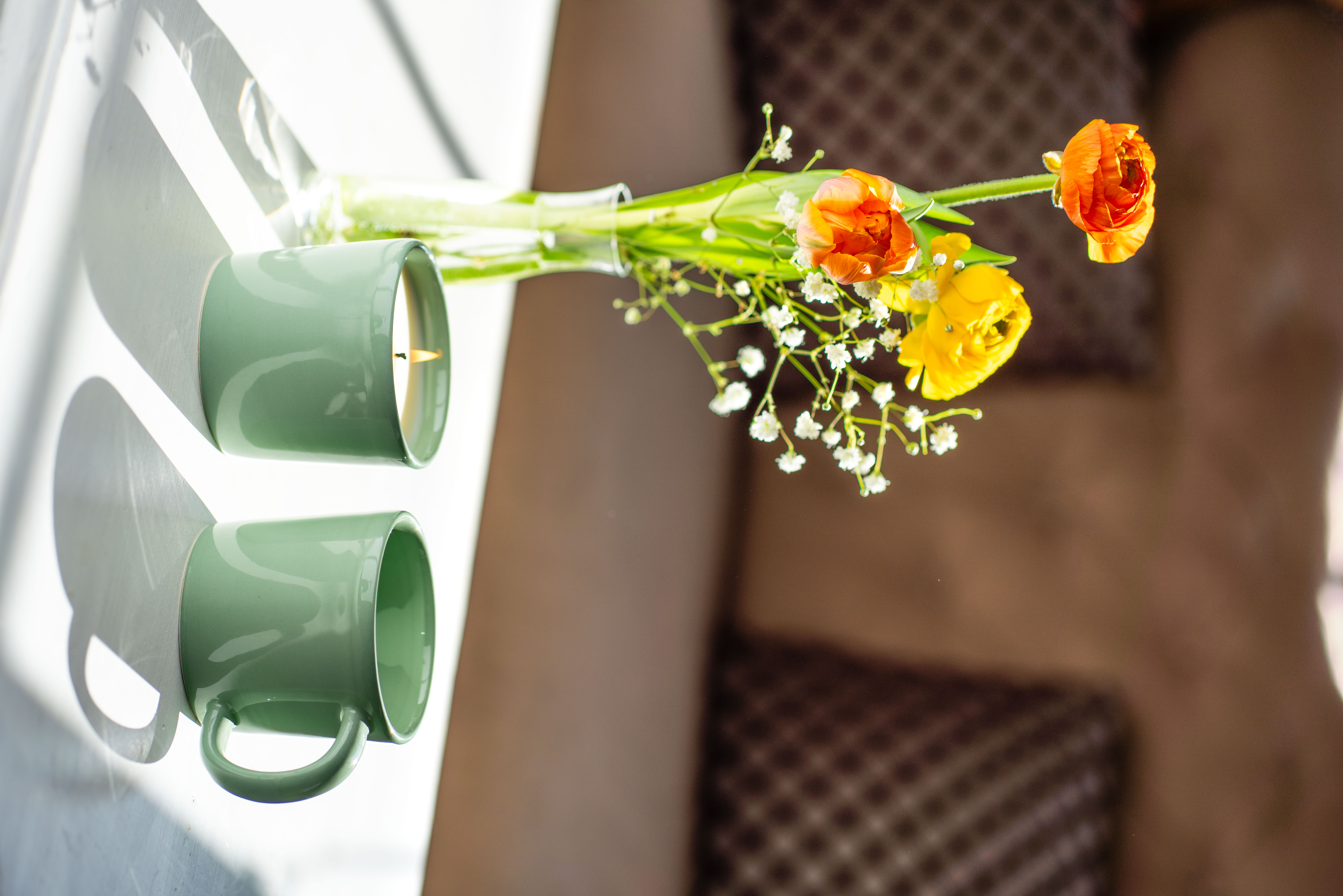 Eine Kerze und eine Kaffeetasse in der Farbe Salbeigrün auf einem Tisch mit drei Frühlingsblumen in einer Vase