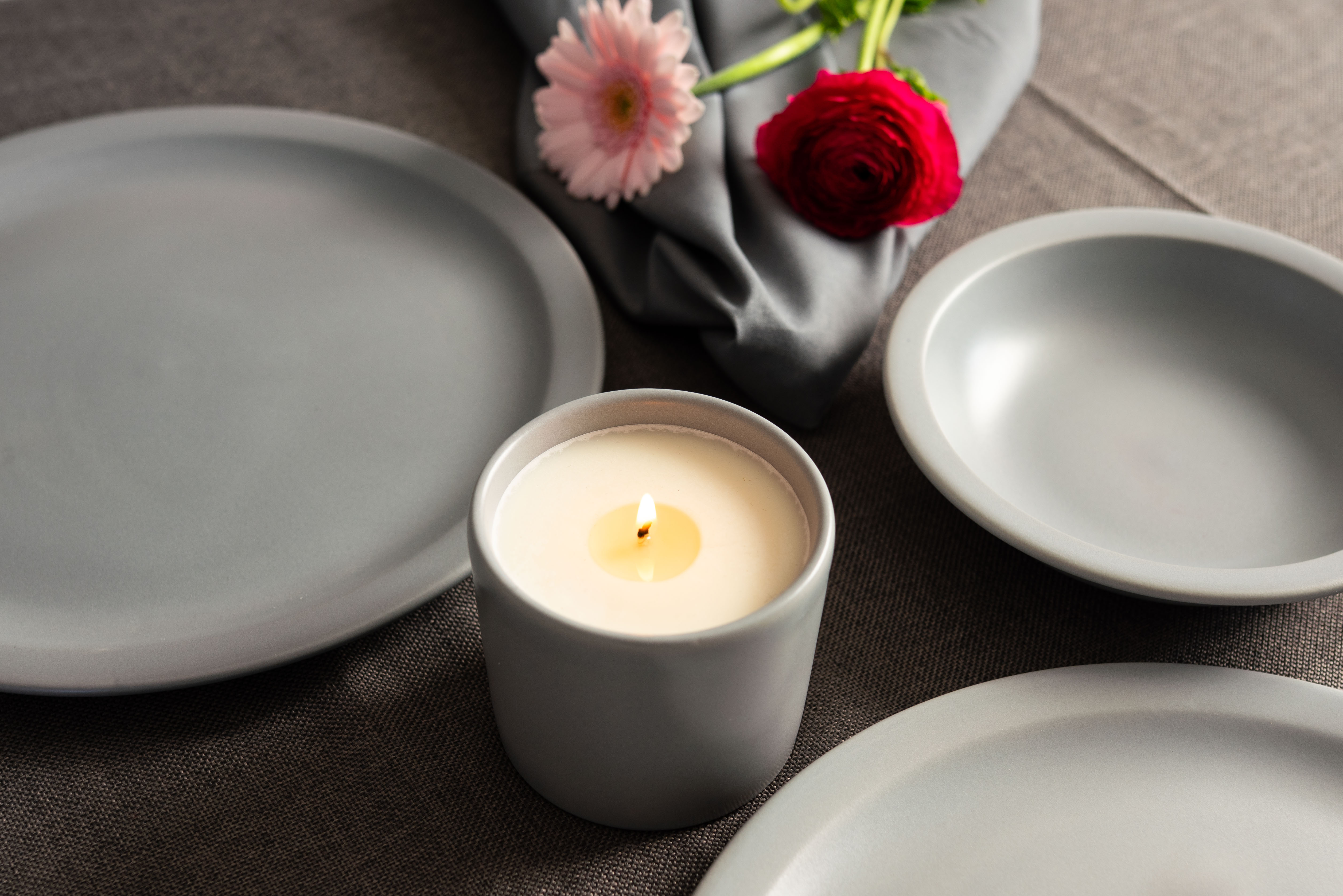 Graue Teller und eine graue Kerze auf einem grauen Tischtuch mit rosafarbenen Blumen daneben