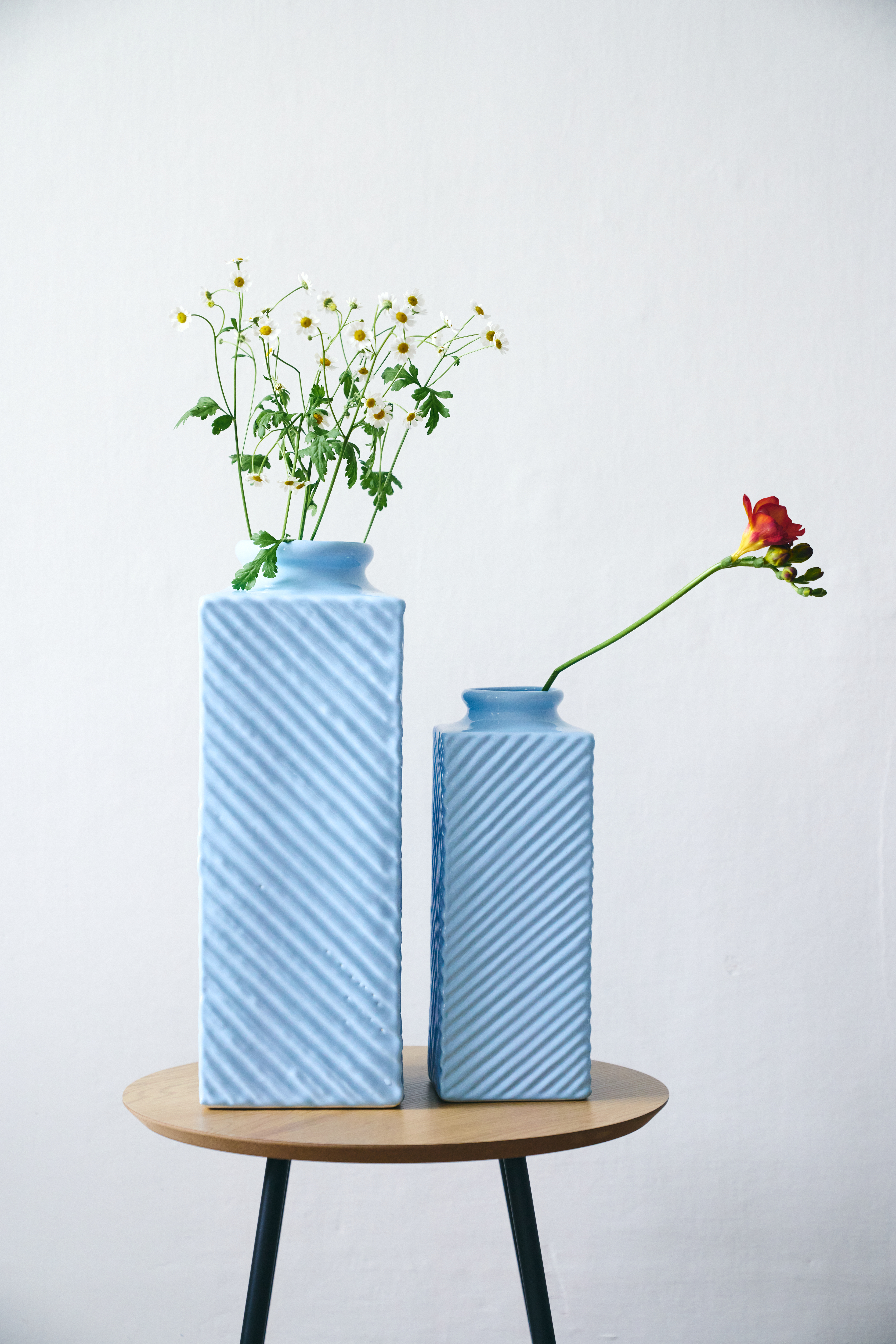 Zwei blaue Vasen mit Blumen stehen vor einer weißen Wand auf einem Holzhocker